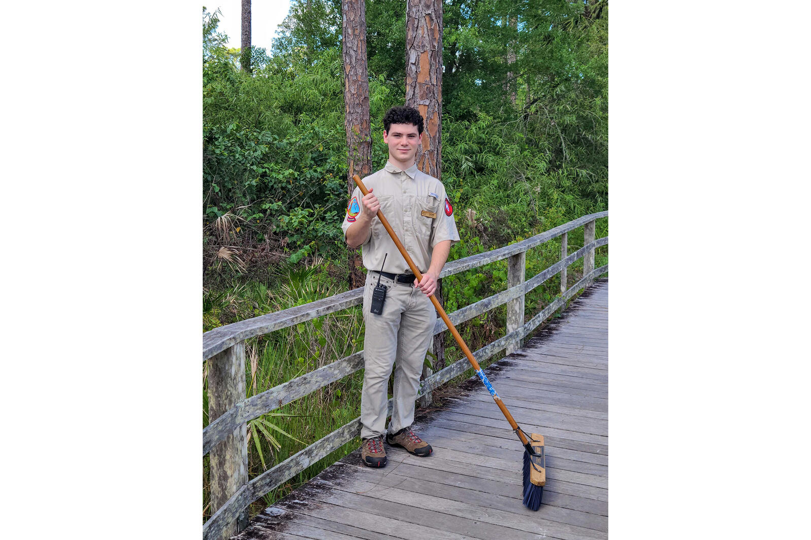 A young man wearing a uniform holds a broom on a boardwalk.