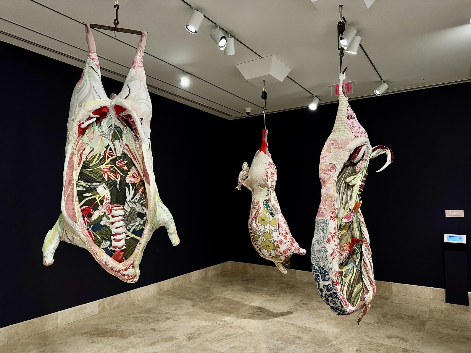 Three fiber art works that resemble hanging cattle carcasses in pastel colors.