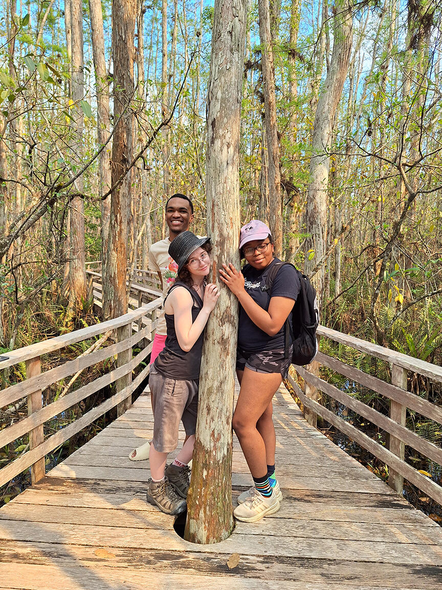 Three people hugging a tree in the middle of a boardwalk.